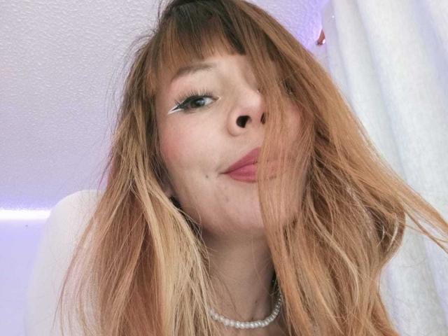 AriiannaHot has long, messy blonde hair, white eyeliner, and a pearl necklace, smiling with pink lipstick, her nose is pierced. Her tits are not visible.