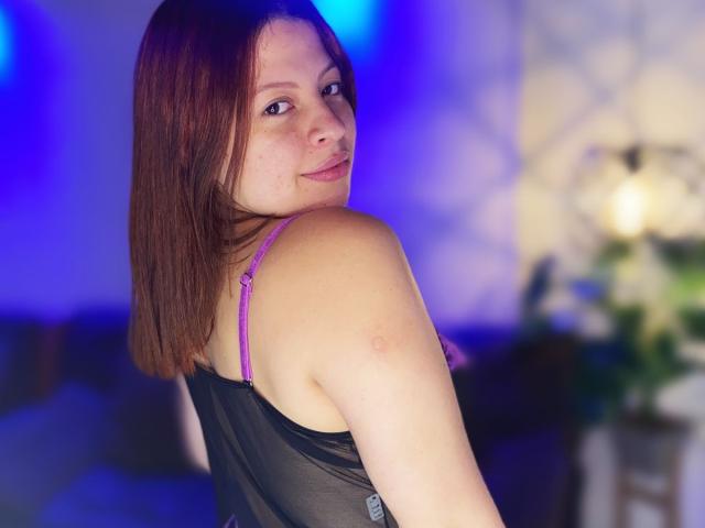ShyVibes with medium brown hair, looking over shoulder, wearing a black sheer top with pink bra straps, slight smile, blue and purple lights in the background.