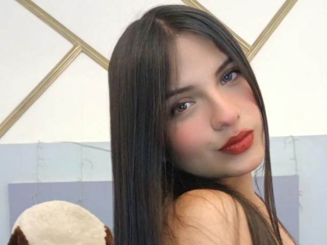 LailaBarker has long, straight black hair, blue eyes, and red lipstick. She's looking over her shoulder with a slight smile. Her tits are partially visible.