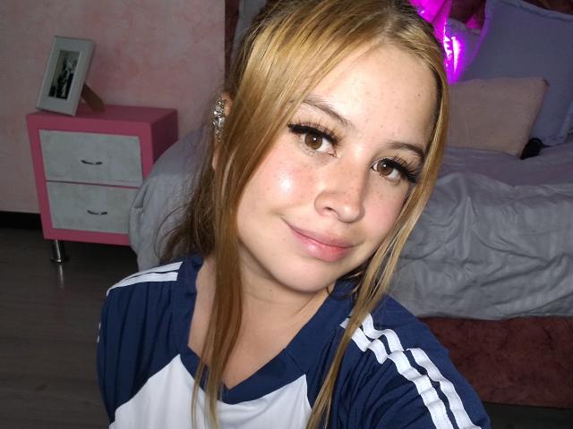 Melani Becker, mit blonden Haaren, trägt ein blau-weißes Sportshirt, lächelt, hat lange Wimpern, helle Haut und kleine Brüste und steht in einem Schlafzimmer mit einem pinken Nachttisch..