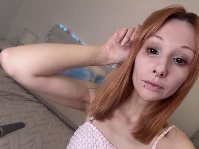 HannaCool has red hair, a nose ring, and lip piercings. She's wearing a pink, textured top, with one arm raised, showing a slim, pale, and cute face.