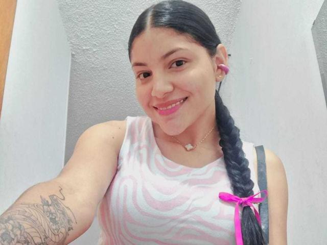 AbriilFoxxy, avec des cheveux noirs tressés, des écouteurs roses, un haut rayé blanc et rose, souriante, montrant un tatouage sur le bras droit, un ruban rose dans les cheveux, du rouge à lèvres rose, portant un collier..