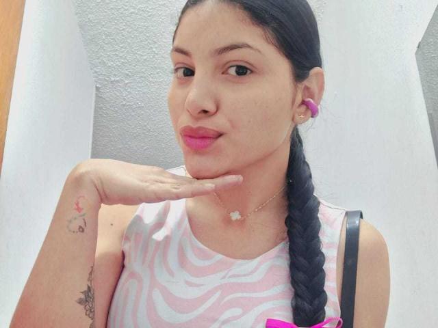 AbriilFoxxy with pink lipstick, braided black hair, zebra-pattern dress, pink earbuds, tattooed hand, and a pink button, posing with her chin up, showing a cute smile.