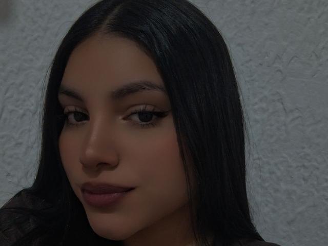 AbriilFoxxy has smooth brown skin, long straight black hair, and dark eyes with winged eyeliner. She's smiling with full pink lips, wearing a dark top.