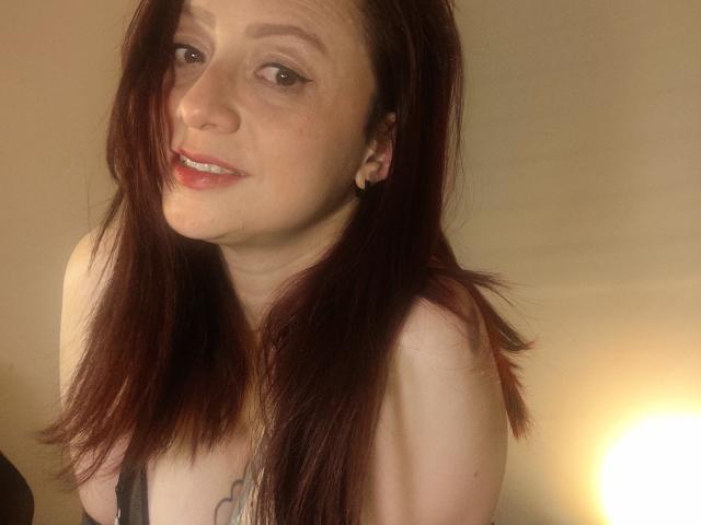 CatalinaMonyque with long red hair, light skin, and a subtle smile, showing small tits, wearing a black strap, and small earrings, in a softly lit indoor setting.