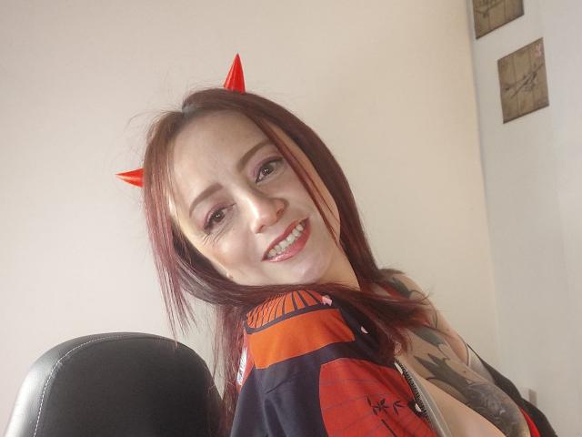 CatalinaMonyque with red devil horns, smiling, showing teeth, red and black sleeveless top, tattoo on right arm, seated in black chair, light skin, red and brown hair.