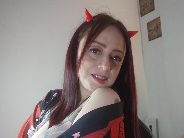 CatalinaMonyque with red devil horns, long red hair, light skin, small nose ring, smiling, wearing red and black kimono, exposing her shoulder, light makeup, slight teeth showing.