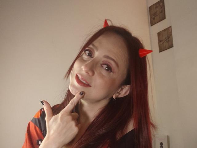 CatalinaMonyque with red devil horns, finger touching her chin, smiling, red lipstick, black nail polish, red and black shirt, pale skin, long red hair, white wall.