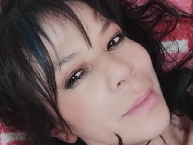 SamaraBellet has dark, wavy hair and brown eyes, with a slight smile on her pink lips. Her skin is fair, and she's lying down on a red and white blanket.