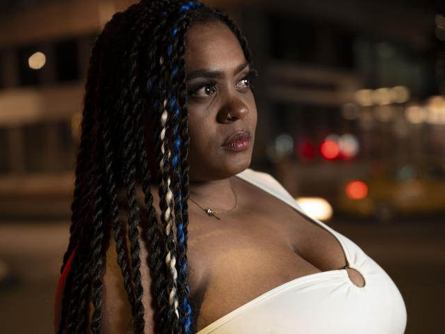 GiorgiaJhenis with large tits, dark brown skin, and blue-black braided hair, wearing a white dress, looking to the side with a serious expression, at night.