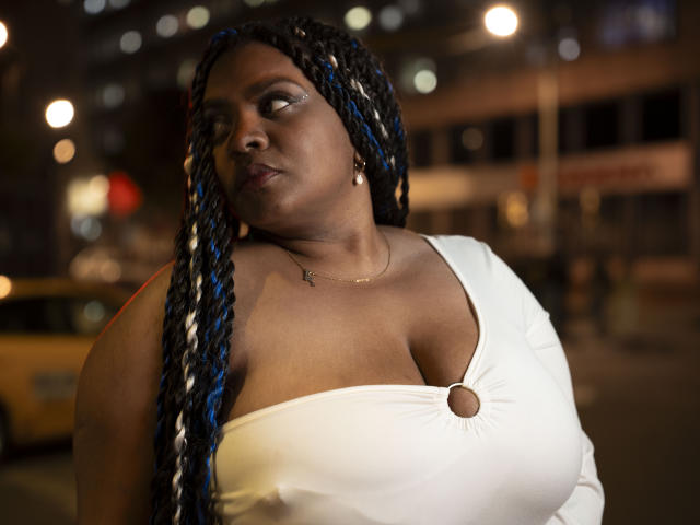 GiorgiaJhenis with large boobs, wearing a white dress with a ring cutout, has blue and black braided hair, and a gold necklace, standing in a nighttime urban setting.