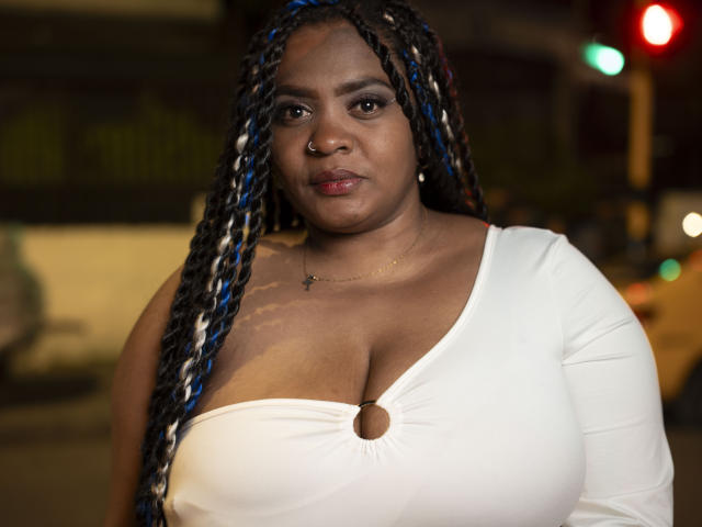 GiorgiaJhenis with big boobs, wearing a white top with a ring, has long blue and black braids, nose ring, and a serious expression, standing at night.