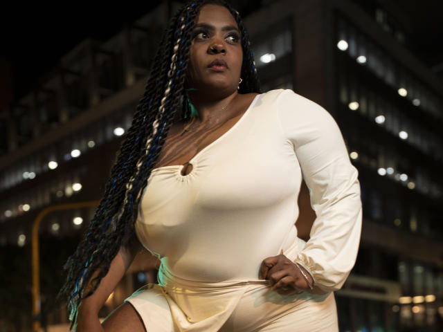 GiorgiaJhenis with large boobs in a white, long-sleeve, one-shoulder dress, standing in a night-time urban setting, hand on hip, looking up with braided hair in blue and white.