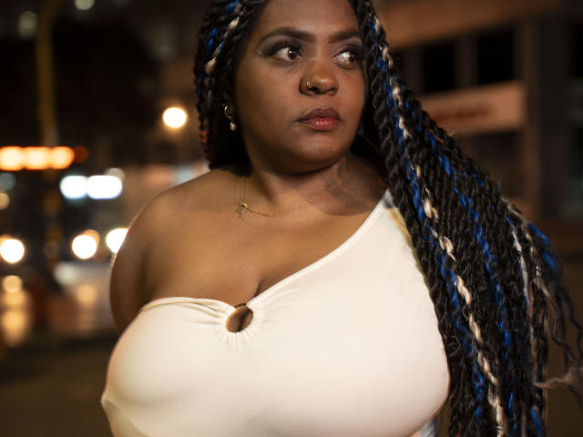 GiorgiaJhenis with large boobs, dark brown skin, and long blue and black braids, wearing a white off-shoulder top, looking to the side, night street background, soft light.
