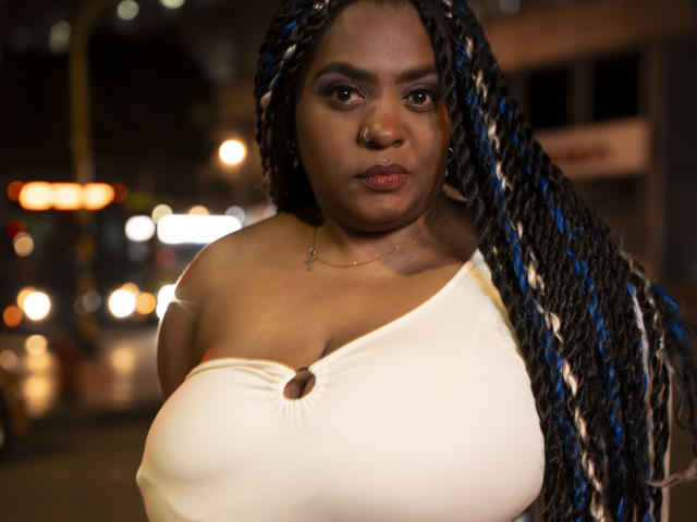 GiorgiaJhenis with dark skin, blue and white braided hair, nose ring, white off-shoulder top, large boobs, serious expression, night setting, city lights in background.
