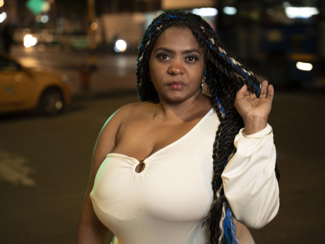 GiorgiaJhenis with large tits, wearing a white off-shoulder top, holding her long blue and black braided hair, standing in a nighttime urban setting.