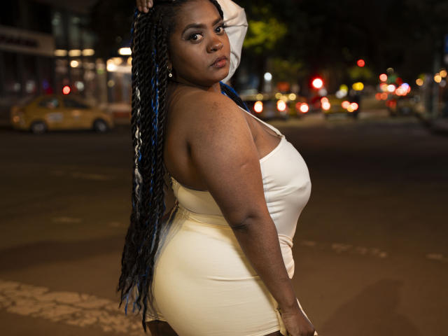 GiorgiaJhenis has dark skin, long blue and black braided hair, and is wearing a tight, cream-colored dress that highlights her big ass and medium-sized tits. She's standing on a street.