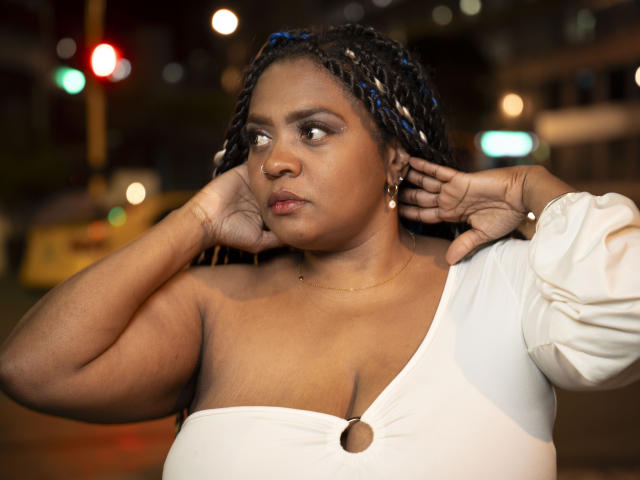 GiorgiaJhenis, dark-skinned, curvy, with blue-braided hair, white off-shoulder top, hands near face, looking to the side, subtle makeup, small nose ring, earrings, night street background.
