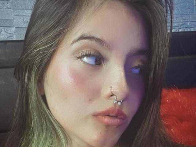 BlairStone has a nose ring with a small silver charm, light brown eyes, and pink lips. Her green-tipped brown hair is slightly messy. She's looking to the side.
