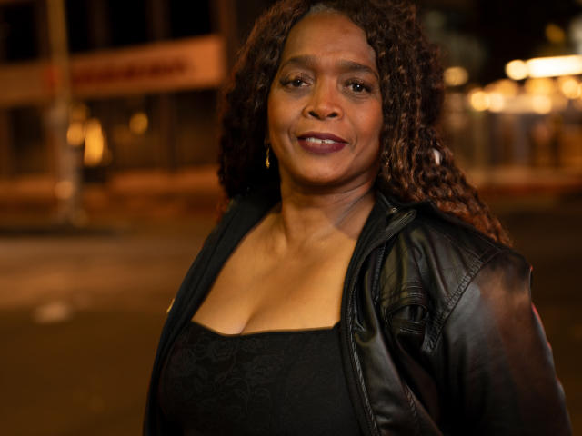 AinaEbonyQueen met krullend bruin haar, gekleed in een zwarte leren jas en een zwarte jurk, die haar middelgrote borsten laat zien, lachend in een stedelijke omgeving 's nachts..
