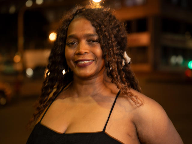 AinaEbonyQueen, con cabello castaño rizado, un top negro con tirantes finos y sonriendo, mostrando sus pechos de tamaño medio y su piel marrón cálida, en un entorno urbano nocturno..