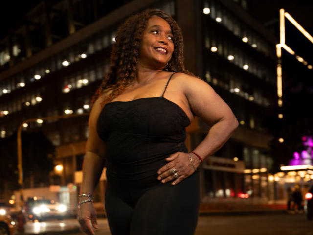 AinaEbonyQueen with curly brown hair, smiling, wearing a black spaghetti strap top, showing her big tits and curvy ass, standing in a city at night, glowing lights behind.