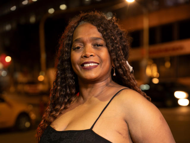 AinaEbonyQueen, una mujer negra de curvas, con cabello castaño rizado, sonriendo, vestida con un vestido negro de encaje, mostrando sus pechos medianos, en un entorno urbano nocturno, con farolas brillantes..
