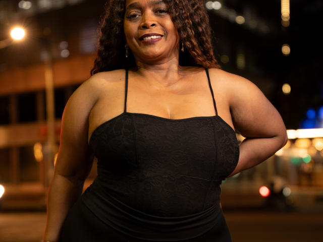 AinaEbonyQueen, curvy black woman with dark curly hair, smiling, wearing a black lace dress with thin straps, showcasing her big tits and soft belly, standing at night.
