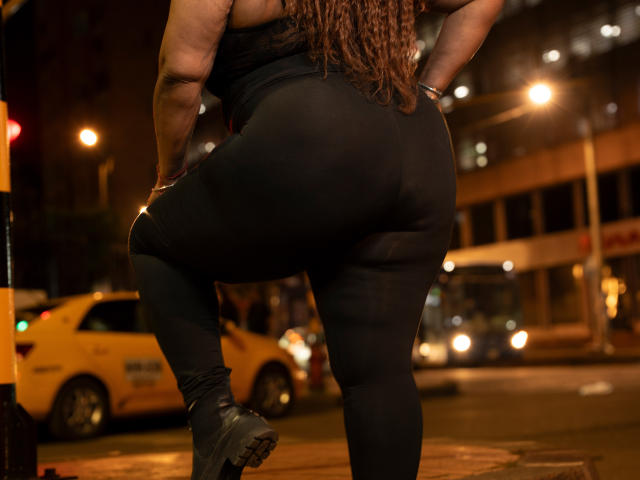 El trasero de AinaEbonyQueen en pantalones negros, ajustados y transparentes, con la zona de los glúteos visible, de pie con un pie sobre un taxi amarillo, llevando botas negras, noche, calle de la ciudad, farolas..