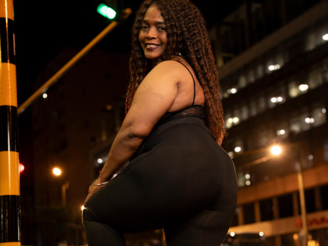 AinaEbonyQueen, con i capelli castano ricci, sorridente, indossa una maglia nera in pizzo e leggings neri aderenti, mostrando il suo grande sedere, in piedi accanto a un palo giallo e nero di notte..