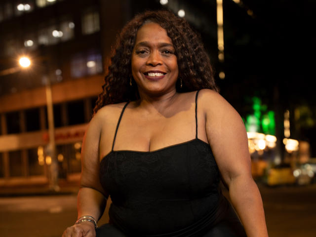 AinaEbonyQueen, una mujer curvilínea con cabello rizado castaño y piel oscura, vestida con un vestido negro con tirantes finos, sonriendo y mostrando sus pechos, en un entorno urbano nocturno..