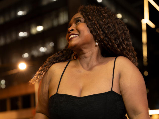 AinaEbonyQueen, une femme noire aux formes généreuses, avec des cheveux bruns bouclés, souriante, portant une robe noire en dentelle avec de fines bretelles, laissant légèrement entrevoir ses seins, dans un décor urbain nocturne..