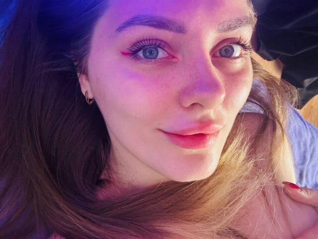TenderAmberr tem olhos azuis, delineador rosa e batom vermelho. Ela está deitada, com cabelos castanhos longos, usando um pequeno brinco de argola e tem esmalte de unha vermelho..