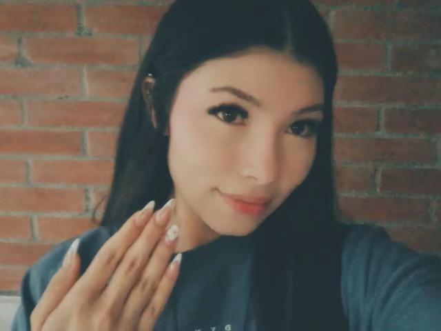 ViolettaLeroy with dark hair, light skin, and long eyelashes, smiling, holding her hand with manicured nails, wearing a blue shirt, looking at the camera.