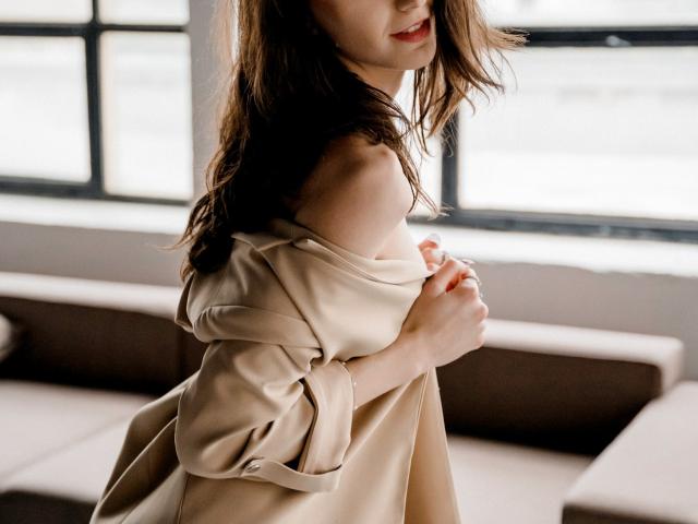 KettyBelly, vestindo um vestido bege com um ombro descoberto, que revela seus pequenos seios, tem cabelo castanho claro, lábios vermelhos e está olhando de lado. Ela está em um quarto iluminado pelo sol, segurando o vestido..