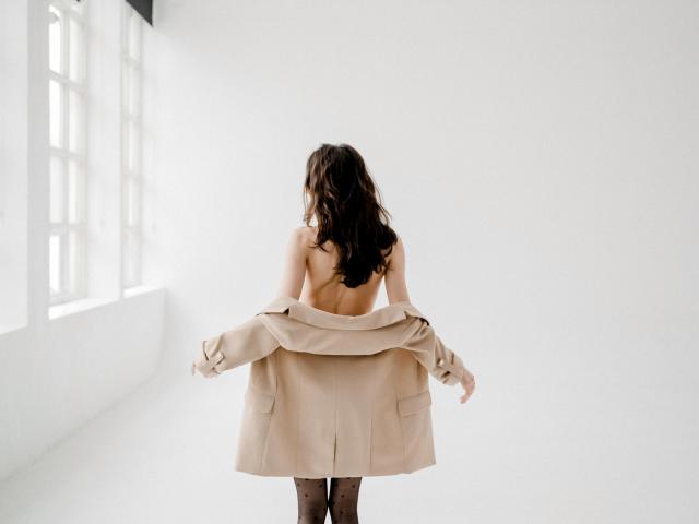 KettyBelly with her back to the camera, topless, pulling off a beige coat, showing her bare ass, wearing black polka dot tights, in a bright, white room.