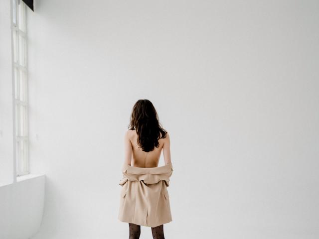 KettyBelly, met haar rug naar de camera, onthult ze haar borsten, met bruin golvend haar, terwijl ze een beige jas over haar billen trekt. Ze staat in een kamer met witte muren en een raam aan de linkerkant..