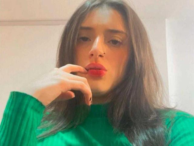 ArielStorm with long brown hair, green sweater, red lipstick, nose ring, and finger near mouth, looking slightly confused. Boobs not visible, focus on face and upper body.