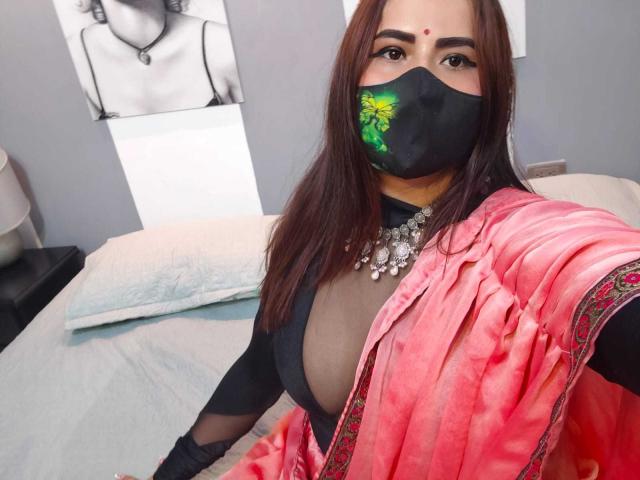 Dalia Dubey, con i capelli castani, una maschera nera con motivo floreale verde, un sari rosa, una maglietta nera che rivela un seno generoso, indossa una collana di diamanti, è seduta su un letto e sta scattando un selfie..