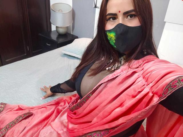 DaliaDubey with dark brown hair, wearing a red saree, black mask, and black bra, showing her tits, sitting on a bed, looking at the camera.