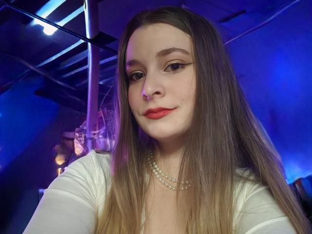 LunaBliss69 has long brown hair, red lipstick, and a pearl necklace, wearing a white top, with a slight smile, in a blue-lit room, showing her face and upper body.