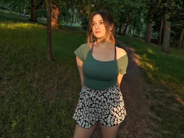 LunaBliss69 standing on a dirt path, green off-shoulder top, black and white floral shorts, hands behind her back, sunlit, grassy park, brown hair, slight smile, natural light.