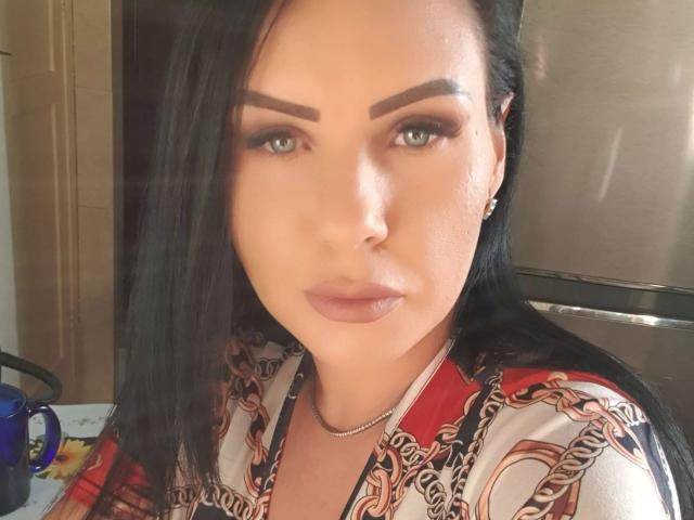 MistressAidaX has dark, straight hair, thick eyebrows, green eyes, and full lips with nude lipstick. She's wearing a red and white patterned top, looking straight at the camera.