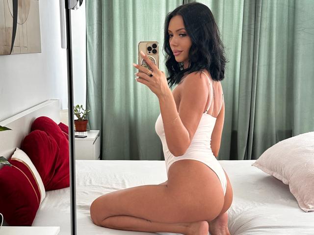 Nadeen, con i capelli neri e ondulati, indossa una body bianca a slip, è inginocchiata sul letto e sta scattando un selfie con il suo iPhone, mostrando i glutei e i piccoli seni..