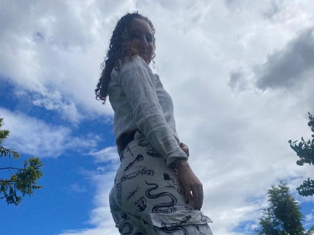 EvaDaMore with curly hair, wearing a gray sweater and white pants with snake patterns, looking down, showing her ass, standing outside under a cloudy blue sky.
