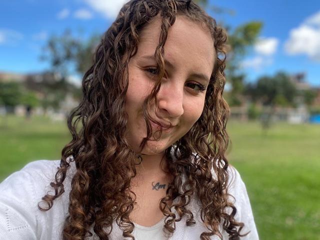 EvaDaMore with curly brown hair, white shirt, slight smile, small tattoo under chin, blue sky, and fluffy clouds in background. Her nose is slightly pinched.