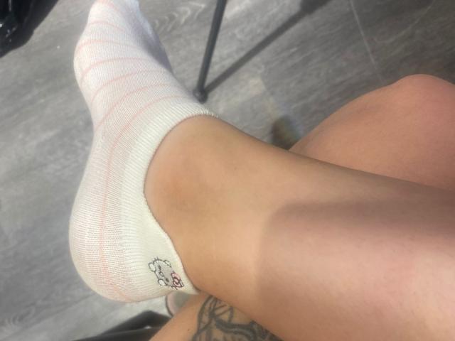 EvaDaMore's foot in a white, striped sock, showing her tan, smooth, and tattooed leg with a small, colorful tattoo near the ankle, on a gray wooden floor.