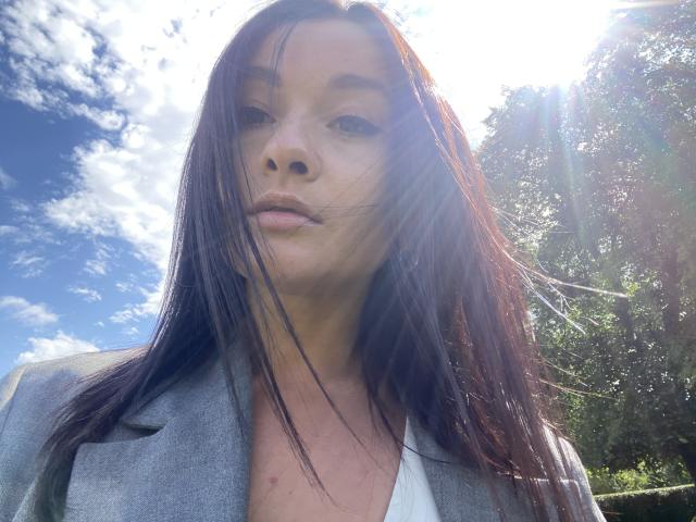 Dionisia has long brown hair, wearing a gray blazer, sunlit face, and slightly parted lips, with a bright blue sky and scattered clouds in the background.