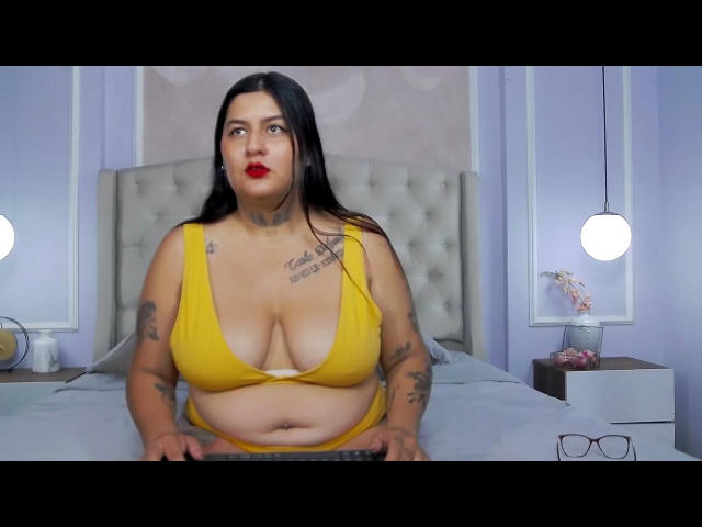 LilyRocks, a curvy Latina woman with black hair, red lipstick, and tattoos, sits on a bed wearing a yellow bra that barely covers her large, saggy tits.