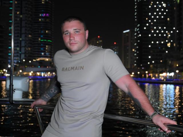OstenFrey, a chubby guy with tattoos, wearing a tight, beige Balmain t-shirt, leaning on a railing, night, city lights reflecting in the water, serious expression.